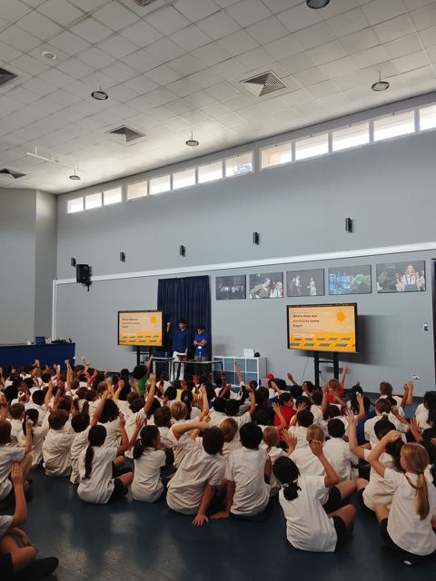 Two Gridovators presenters stand between two screens that ask where our electricity comes from, while a school hall full of students raise their hands.