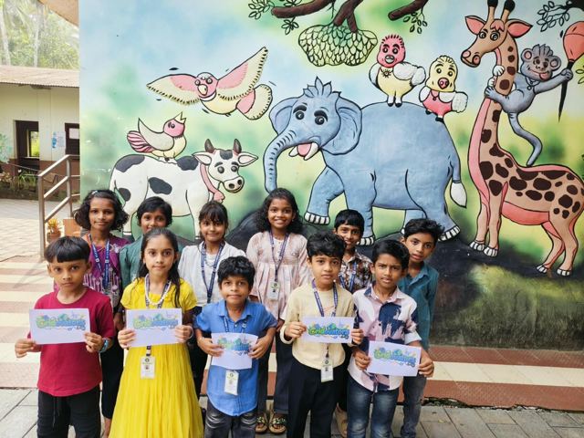 Students at GLPS Nediyanad West stand in front of a painted animal mural, some holding up sheets printed with the Gridovators logo.