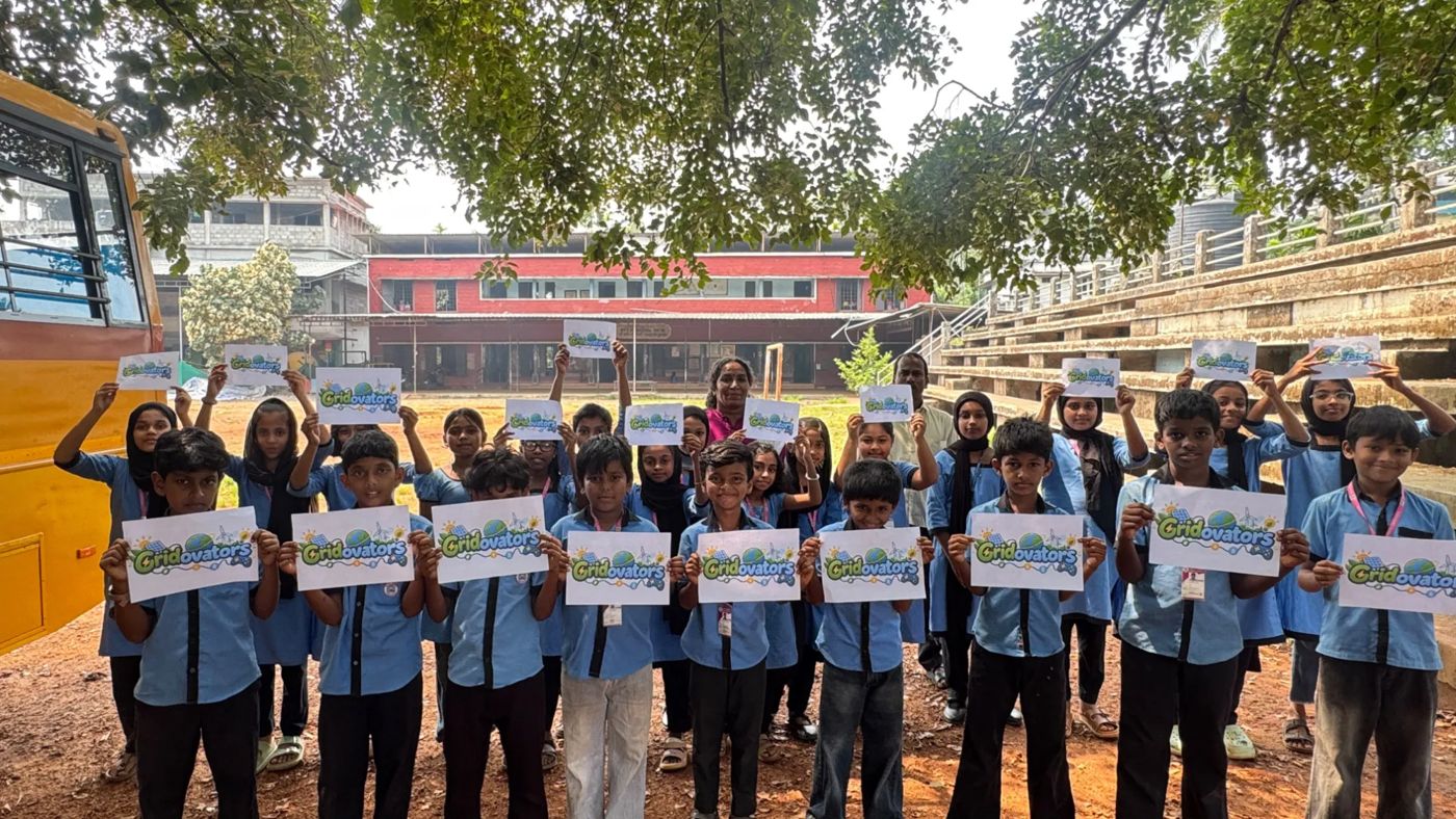 A large group of students in blue school uniforms holds up Gridovators logo sheets in a school yard, with two teachers standing among them.